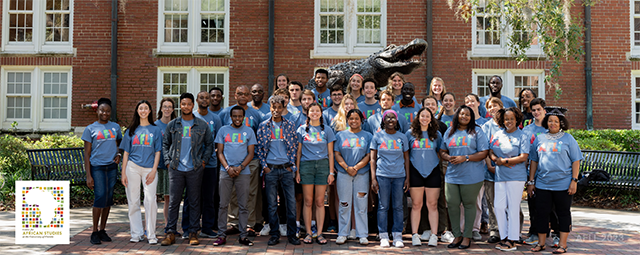 A large group of students pose in front of the On Top of the World sculpture at Heavener Hall, UF. The students are smiling at the camera. They are all wearing light-blue T-shirts that display the multicolored African Languages Institute (AFLI) logo.