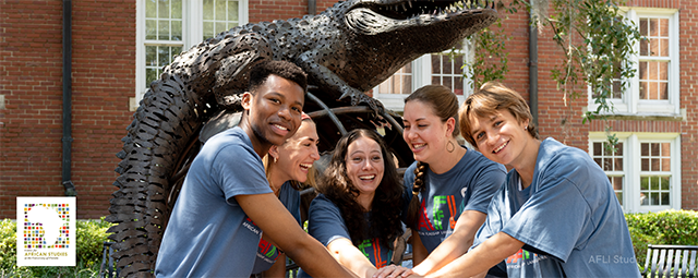 A small group of students pose in front of the On Top of the World sculpture at Heavener Hall, UF. The students are smiling at the camera, each of them placing one hand in the center, on top of each others' hands. The ﻿On Top of the World sculpture at Heavener Hall, UF depicts a Gator on top of a globe.