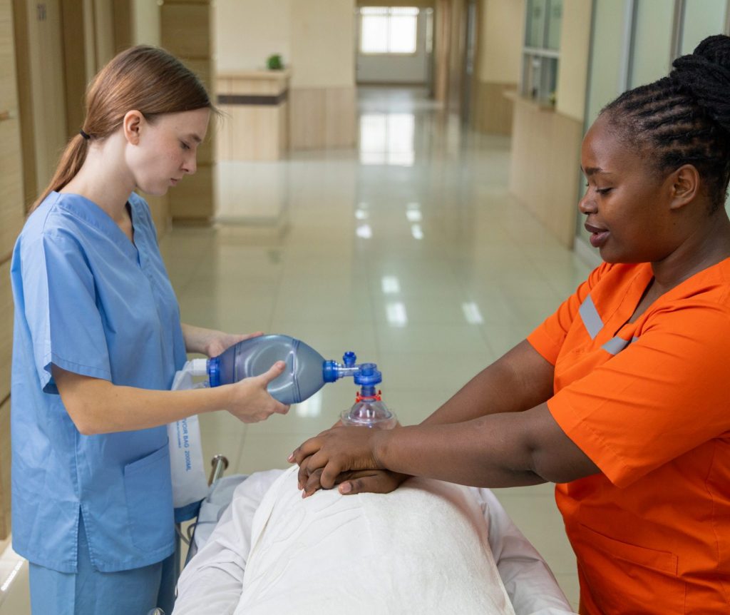 Medical emergency with patient receiving oxygen: Close-up of unconscious man on stretcher receiving manual ventilation from medical staff using an oxygen mask in hospital hallway.